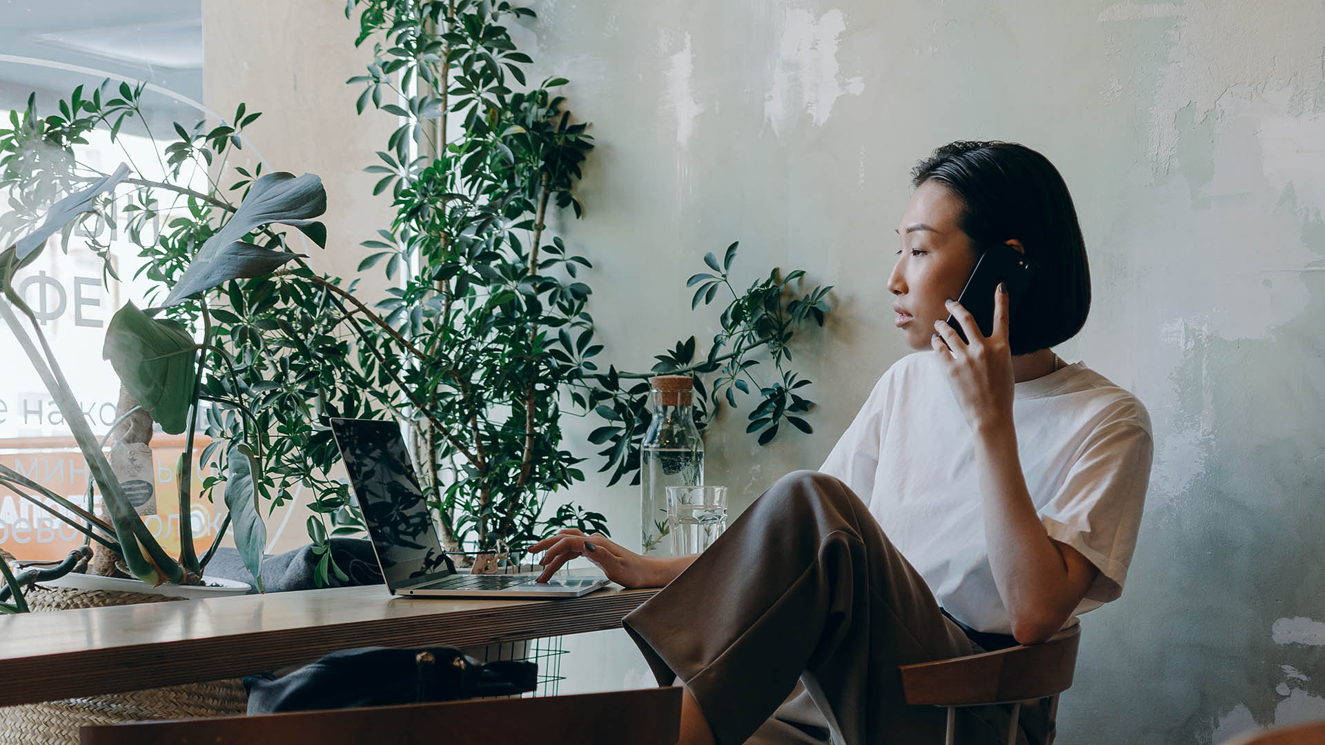 Person in white shirt and brown pants talking on phone while seated at table with computer.