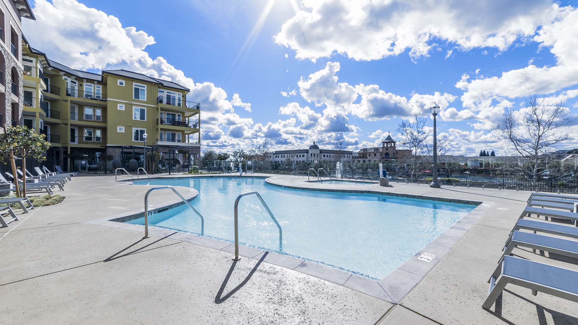 Pool area with lounge chairs, spa, and metal fence overlooked by apartment balconies.